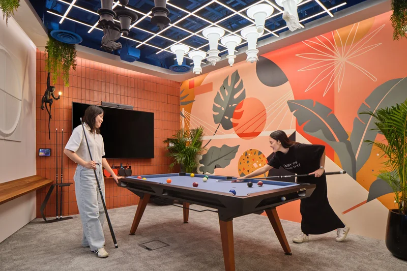 Two women playing pool in a vibrant office recreation room with a tropical leaf mural in orange and green, blue grid ceiling, and hanging plants.